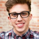 Portrait of a smiling young man wearing eyeglasses and plaid shirt against a brick wall background.